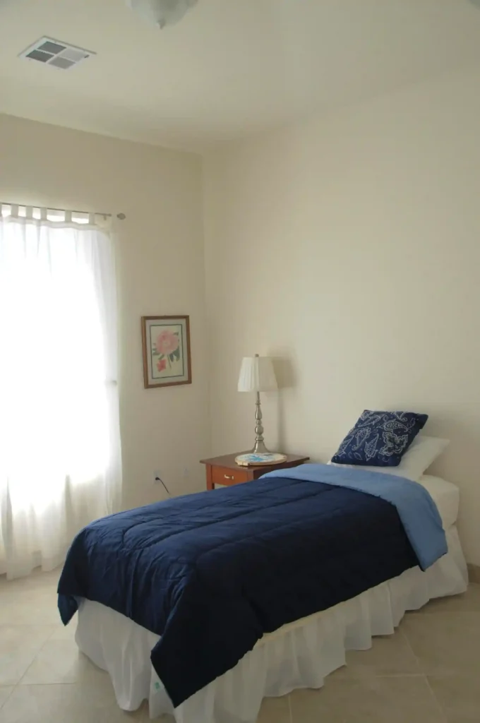 In this image you can see, A bed with a blanket on it, a lamp with its table and wall with a light coming.